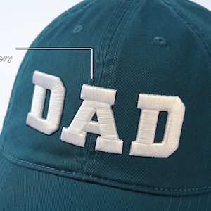 May include: A teal blue baseball cap with white 3D embroidery that reads "DAD".