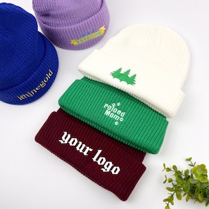 May include: Five knitted beanies in different colors with embroidered designs. The beanies are stacked on top of each other. The designs include "Freedom", "Polished Mom", "Your Logo", and a pine tree.