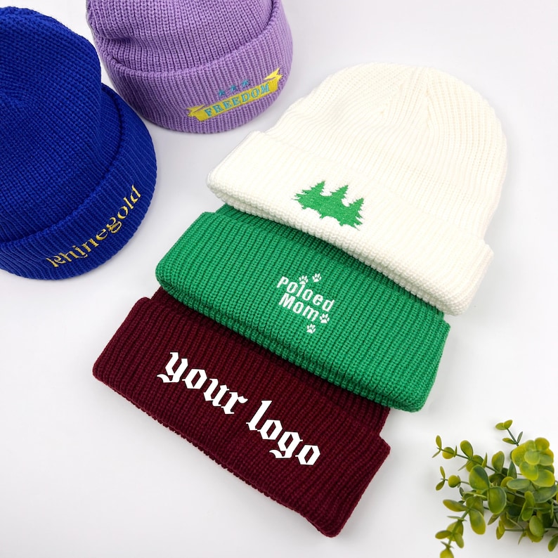 May include: Five knitted beanies in different colors with embroidered designs. The beanies are stacked on top of each other. The designs include "Freedom", "Polished Mom", "Your Logo", and a pine tree.