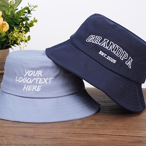 May include: Two bucket hats, one light blue and one navy blue, are displayed on a wooden surface. The light blue hat has the text "YOUR LOGO/TEXT HERE" embroidered in white. The navy blue hat has the word "GRANDPA" and "EST. 2025" embroidered in white.