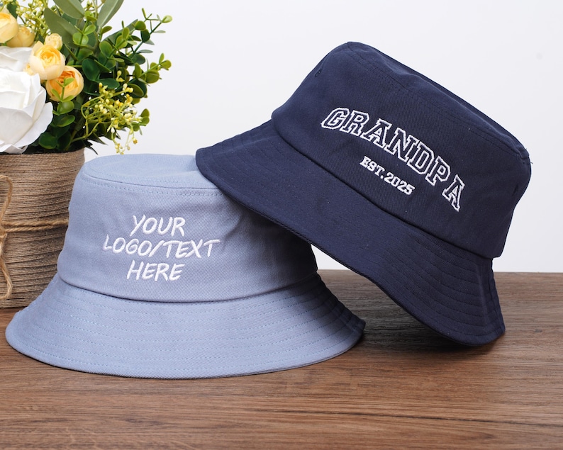 May include: Two bucket hats, one light blue and one navy blue, are displayed on a wooden surface. The light blue hat has the text "YOUR LOGO/TEXT HERE" embroidered in white. The navy blue hat has the word "GRANDPA" and "EST. 2025" embroidered in white.