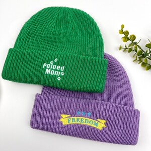 May include: Two knitted beanies, one green with "Pawed Mom" embroidered in white, and one purple with "FREEDOM" embroidered in yellow on a blue banner.