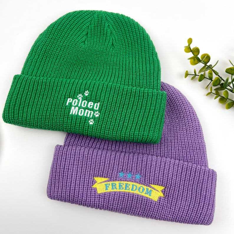 May include: Two knitted beanies, one green with "Pawed Mom" embroidered in white, and one purple with "FREEDOM" embroidered in yellow on a blue banner.