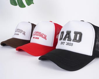Könnte beinhalten: Drei Trucker-Caps mit weißen Fronten und Mesh-Rücken. Eine hat einen braunen Schirm und das Wort "GRANDPA" in Weiß gestickt, eine andere hat einen roten Schirm mit "GRANDMA" in Rot, und die dritte hat einen schwarzen Schirm mit "DAD" in Grau. Alle Caps haben "EST 2025".