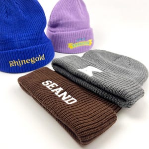 May include: Four knitted beanies in different colors with embroidered text. The beanies are blue, purple, gray, and brown. The text on the beanies reads "Rhinegold", "FREEDOM", "R", and "SEAND".