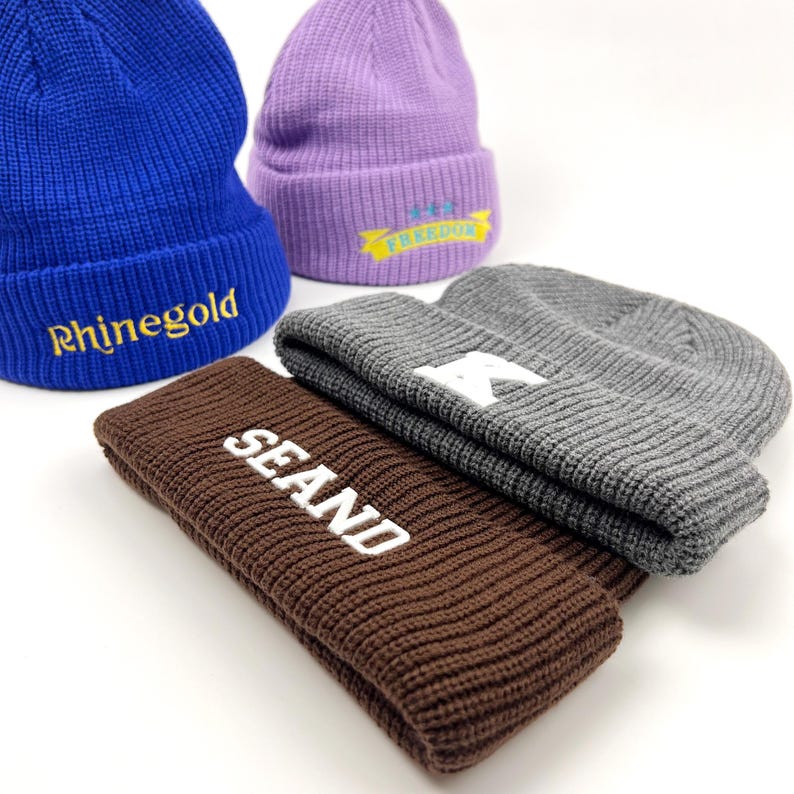 May include: Four knitted beanies in different colors with embroidered text. The beanies are blue, purple, gray, and brown. The text on the beanies reads "Rhinegold", "FREEDOM", "R", and "SEAND".