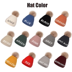 May include: A collection of thirteen ribbed knit beanies with faux fur pom-poms, displayed in various colors including white, blue, black, orange, beige, green, pink, grey, navy blue, yellow, red, light grey, and burgundy, each labeled with its color.