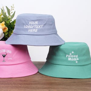 May include: Three pastel-coloured bucket hats: light blue, pink, and teal. The light blue hat has the text "YOUR LOGO/TEXT HERE". The pink hat has a martini glass graphic and the text "CUSTOM LOGO EST 1998". The teal hat says "Poloed Mom".