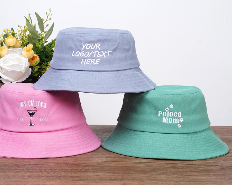 May include: Three pastel-coloured bucket hats: light blue, pink, and teal. The light blue hat has the text "YOUR LOGO/TEXT HERE". The pink hat has a martini glass graphic and the text "CUSTOM LOGO EST 1998". The teal hat says "Poloed Mom".