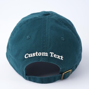 K&ouml;nnte beinhalten: Eine marineblaue Baseballkappe mit verstellbarem Riemen und wei&szlig; gesticktem Text, der "Custom Text" lautet.