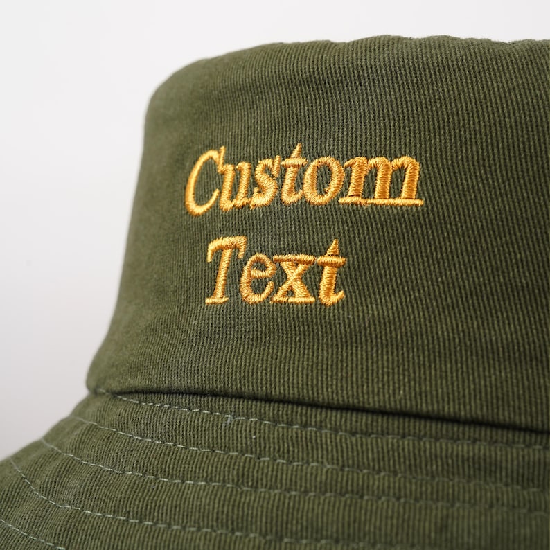Może przedstawiać: Zielony kapelusz typu bucket w kolorze oliwkowym z wyhaftowanym złotym napisem "Custom Text". Kapelusz wykonany jest z teksturowanej tkaniny i ma klasyczny, swobodny styl.