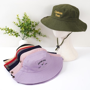 Könnte beinhalten: Eine Auswahl an Fischerhüten in verschiedenen Farben, darunter Olivgrün, Rosa, Marineblau und Beige. Ein Hut, der auf einem Schaufensterkopf präsentiert wird, trägt den Text "Custom Text". Ein anderer Hut hat ein individuelles Logo und "EST 1996".
