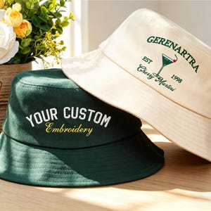 May include: Two bucket hats, one cream and one dark green, are displayed. The cream hat has a green embroidered cocktail design with the text "GERENARTRA EST 1998 Cherry Martini." The green hat features the text "YOUR CUSTOM Embroidery" in white and gold.