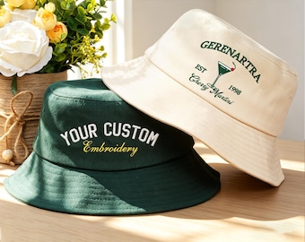 May include: Two bucket hats are shown, one cream and one dark green. The cream hat has a green embroidered cocktail design with the text "GERENARTRA EST 1998 Cherry Martini." The green hat features the text "YOUR CUSTOM Embroidery" in white and gold.