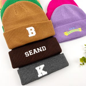 May include: A stack of five knitted beanies in various colors, including brown, purple, gray, and black. The beanies have different embroidered designs, including the letters "B", "K", and "SEAND", and the word "FREEDOM" with stars.