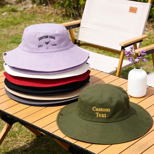 Może przedstawiać: Stos kapeluszy typu bucket w r&oacute;żnych kolorach, w tym lawendowym, białym, czerwonym, czarnym i oliwkowym. Lawendowy kapelusz ma wyhaftowane "CUSTOM LOGO" i grafikę koktajlu. Oliwkowy kapelusz ma napis "Custom Text" w kolorze ż&oacute;łtym.