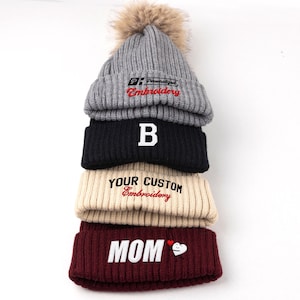 May include: A stack of four ribbed knit beanies. The top grey beanie has a faux fur pom-pom and text reading Personalized Embroidery. The black beanie features a white letter B. The beige beanie reads YOUR CUSTOM Embroidery. The maroon beanie reads MOM.