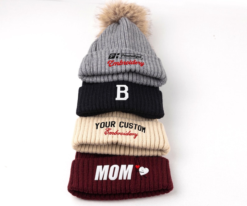 May include: A stack of four ribbed knit beanies. The top grey beanie has a faux fur pom-pom and text reading Personalized Embroidery. The black beanie features a white letter B. The beige beanie reads YOUR CUSTOM Embroidery. The maroon beanie reads MOM.