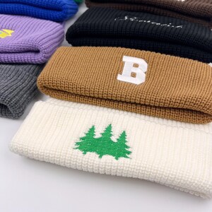 May include: A stack of knitted beanies in various colors, including brown, white, black, purple, and gray. The brown beanie has a white letter "B" embroidered on it. The white beanie has a green embroidered design of three pine trees.