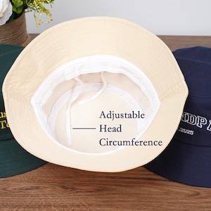 May include: Three bucket hats are displayed, with the beige hat in the center showing the adjustable head circumference. The other hats are dark green and navy blue. The dark green hat has gold embroidery, and the navy blue hat has white embroidery.