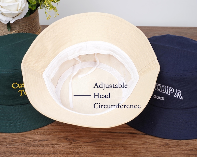 May include: Three bucket hats are displayed, with the beige hat in the center showing the adjustable head circumference. The other hats are dark green and navy blue. The dark green hat has gold embroidery, and the navy blue hat has white embroidery.