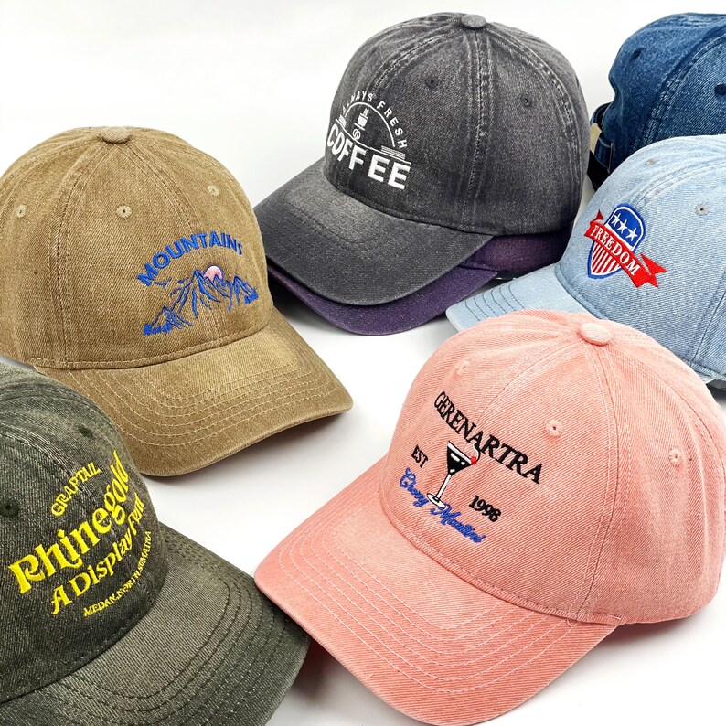 May include: A selection of baseball caps in shades like brown, grey, pink, and blue. Each cap is embroidered with designs and text, including "Mountains," "Coffee," and "Freedom." The caps have a vintage, worn appearance.
