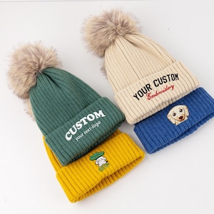 May include: A collection of four ribbed knit beanies featuring large faux fur pom-poms. The hats come in green, yellow, cream, and blue. The green hat says CUSTOM your text-logo, the cream hat says YOUR CUSTOM Embroidery, and the yellow hat has a small graphic.