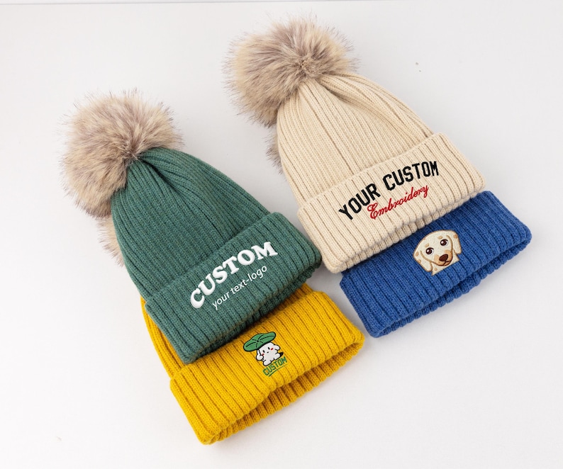 May include: A collection of four ribbed knit beanies featuring large faux fur pom-poms. The hats come in green, yellow, cream, and blue. The green hat says CUSTOM your text-logo, the cream hat says YOUR CUSTOM Embroidery, and the yellow hat has a small graphic.