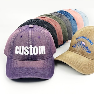 May include: A selection of baseball caps in shades like purple, black, and tan. The purple cap displays the word "custom" in white. The tan cap has a mountain and sun design embroidered on it.