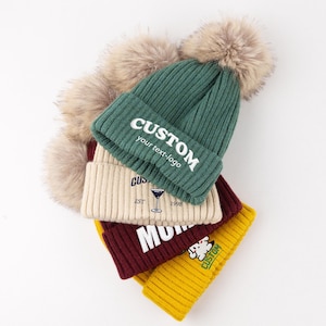 May include: A stack of four ribbed knit beanies in dark green, cream, maroon, and yellow, each topped with a large beige faux fur pom-pom. The green beanie features the white text CUSTOM your text-logo, while the others display various graphics.
