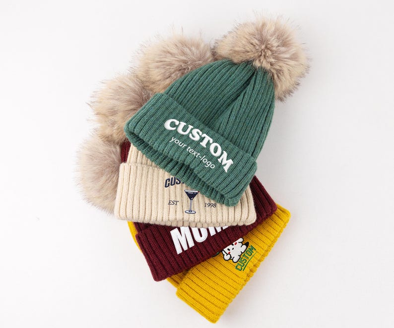 May include: A stack of four ribbed knit beanies in dark green, cream, maroon, and yellow, each topped with a large beige faux fur pom-pom. The green beanie features the white text CUSTOM your text-logo, while the others display various graphics.