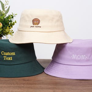 May include: Three bucket hats in cream, dark green, and lavender. The cream hat has a dog embroidery with "pet name" text. The dark green hat has "Custom Text" embroidered in gold. The lavender hat has "mom" embroidered.