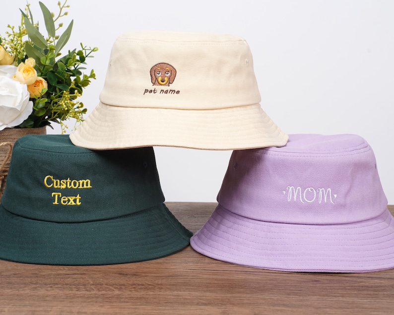 May include: Three bucket hats in cream, dark green, and lavender. The cream hat has a dog embroidery with "pet name" text. The dark green hat has "Custom Text" embroidered in gold. The lavender hat has "mom" embroidered.