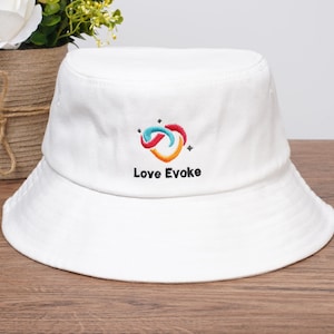 May include: White bucket hat with a colourful embroidered logo and the text "Love Evoke". The hat is made of a soft material and has a wide brim. The logo features a swirl design in shades of blue, red, and orange.