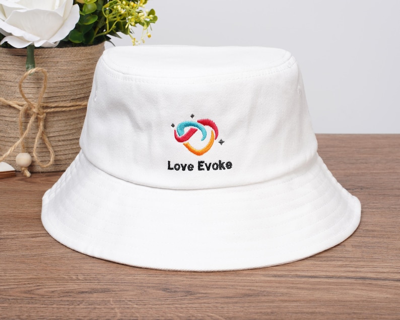 May include: White bucket hat with a colourful embroidered logo and the text "Love Evoke". The hat is made of a soft material and has a wide brim. The logo features a swirl design in shades of blue, red, and orange.