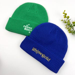 May include: Two knitted beanies, one green with white text that says "Pawed Mom" and the other blue with gold text that says "Rhinegold".
