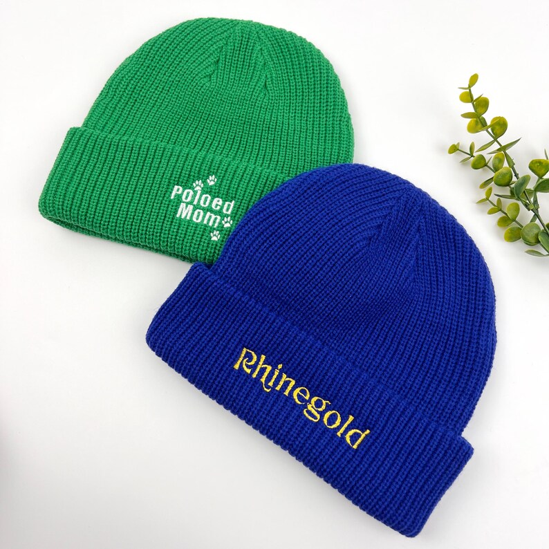 May include: Two knitted beanies, one green with white text that says "Pawed Mom" and the other blue with gold text that says "Rhinegold".