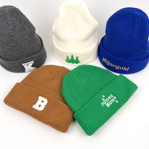May include: Five knit beanies in different colors with embroidered designs. The beanies are gray, white, blue, brown, and green. The designs include a letter K, a tree, the word "shinegold", a letter B, and the words "Pawed Mom".