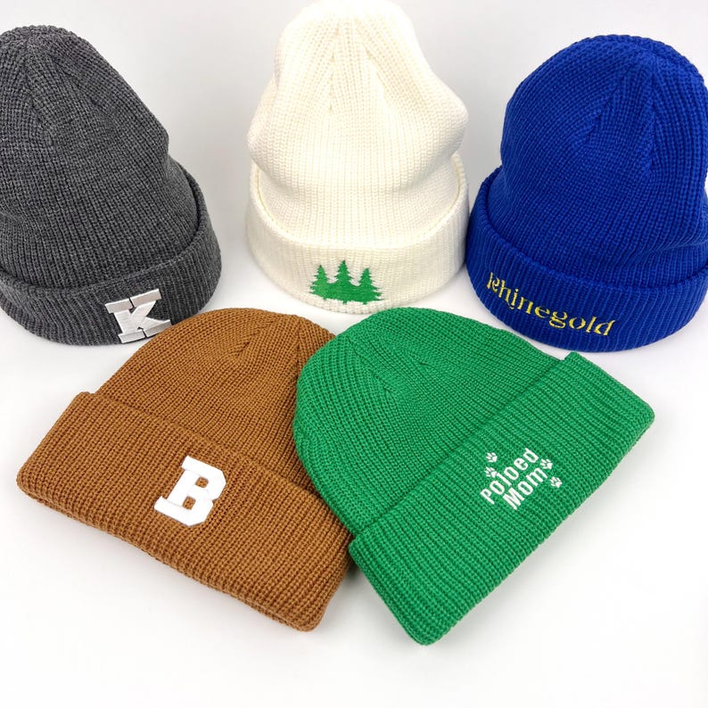 May include: Five knit beanies in different colors with embroidered designs. The beanies are gray, white, blue, brown, and green. The designs include a letter K, a tree, the word "shinegold", a letter B, and the words "Pawed Mom".