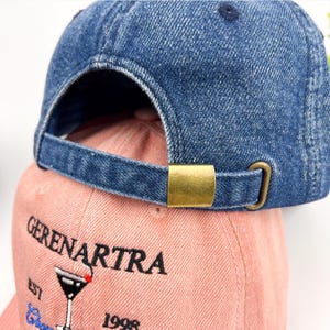 May include: Two baseball caps, one denim blue and one pink. The blue cap's brim is visible, with a brass-coloured metal clasp. The pink cap has the text "GERENARTRA" and a cocktail glass design embroidered on the front.