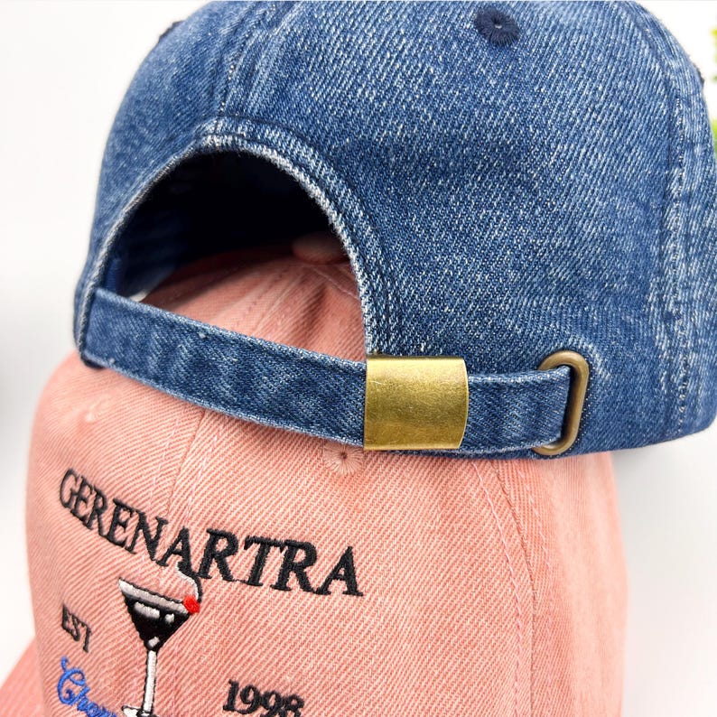 May include: Two baseball caps, one denim blue and one pink. The blue cap's brim is visible, with a brass-coloured metal clasp. The pink cap has the text "GERENARTRA" and a cocktail glass design embroidered on the front.