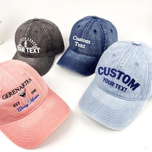 Może przedstawiać: Cztery czapki bejsbolowe w różnych kolorach: ciemnoszarym, dżinsowym niebieskim i jasnoróżowym. Każda czapka ma haftowany tekst, w tym "Custom Text", "Always Fresh" i grafikę koktajlu. Czapki są wykonane z miękkiej, pranej tkaniny.