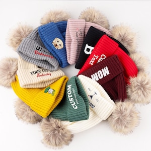 May include: A collection of ten ribbed knit beanies arranged in a circular pattern, each featuring a large faux fur pom-pom on top. The beanies come in various colors including gray, blue, pink, black, red, maroon, yellow, green, and white.
