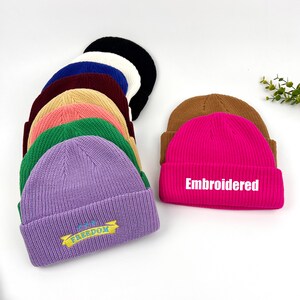 May include: A collection of ten knitted beanies in various colors, including purple, pink, green, brown, and white. Some beanies have embroidered designs, including "Embroidered" and "The Freedom".