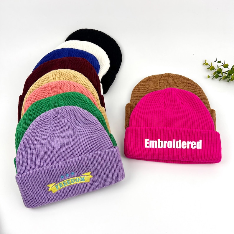 May include: A collection of ten knitted beanies in various colors, including purple, pink, green, brown, and white. Some beanies have embroidered designs, including "Embroidered" and "The Freedom".
