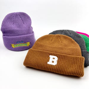 May include: Five knitted beanies in different colors. One beanie is purple with the text "F.A.M. FREEDOM" embroidered on it. Another beanie is brown with a white letter "B" embroidered on it. The other beanies are gray, pink, and green.