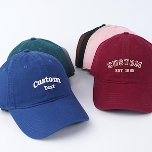 K&ouml;nnte beinhalten: Eine Gruppe von sechs Baseballkappen in verschiedenen Farben, darunter Blau, Rot, Gr&uuml;n, Pink und Schwarz. Die Kappen sind alle aus Baumwolle und haben ein klassisches, unstrukturiertes Design. Die blaue Kappe hat den Text "Custom Text" auf der Vorderseite gestickt, w&auml;hrend die rote Kappe den Text "CUSTOM EST 1985" auf der Vorderseite gestickt hat.