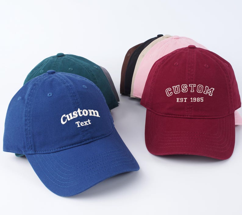 K&ouml;nnte beinhalten: Eine Gruppe von sechs Baseballkappen in verschiedenen Farben, darunter Blau, Rot, Gr&uuml;n, Pink und Schwarz. Die Kappen sind alle aus Baumwolle und haben ein klassisches, unstrukturiertes Design. Die blaue Kappe hat den Text "Custom Text" auf der Vorderseite gestickt, w&auml;hrend die rote Kappe den Text "CUSTOM EST 1985" auf der Vorderseite gestickt hat.