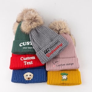 May include: A collection of five ribbed knit beanies with faux fur pom-poms in green, red, blue, gray, pink, and yellow. The beanies feature embroidered text and graphics including 'CUSTOM your text-here', 'Custom Text', a dog face, 'Personalized Embroidery', 'custom design', and a small character under a leaf.