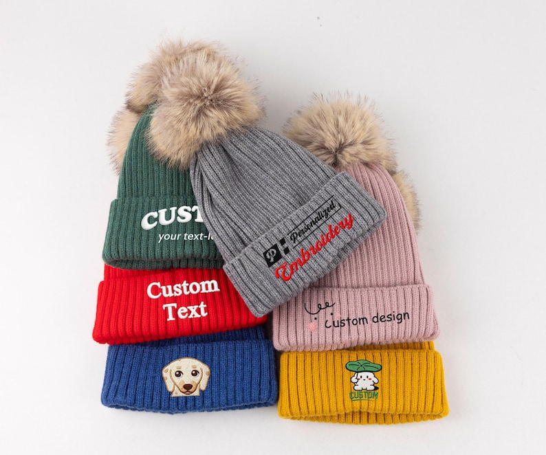 May include: A collection of five ribbed knit beanies with faux fur pom-poms in green, red, blue, gray, pink, and yellow. The beanies feature embroidered text and graphics including 'CUSTOM your text-here', 'Custom Text', a dog face, 'Personalized Embroidery', 'custom design', and a small character under a leaf.
